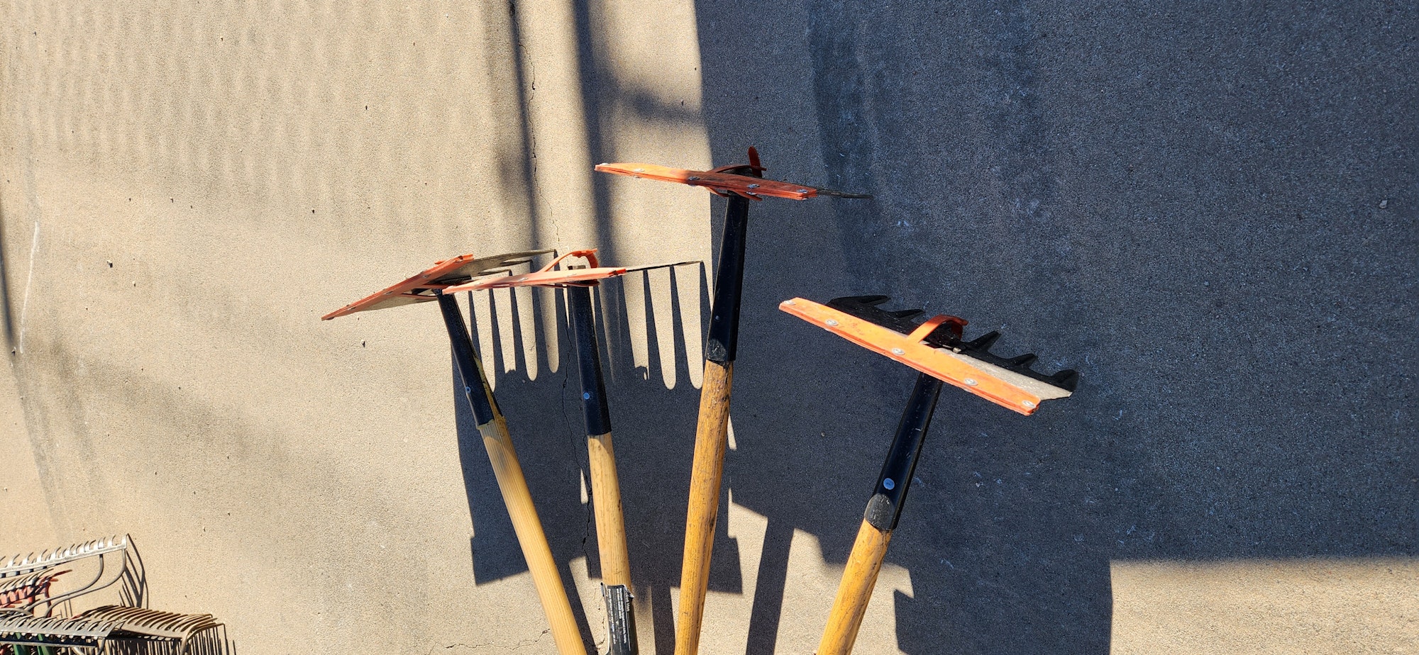 Gardening tools with wooden handles on an asphalt surface beside sand.