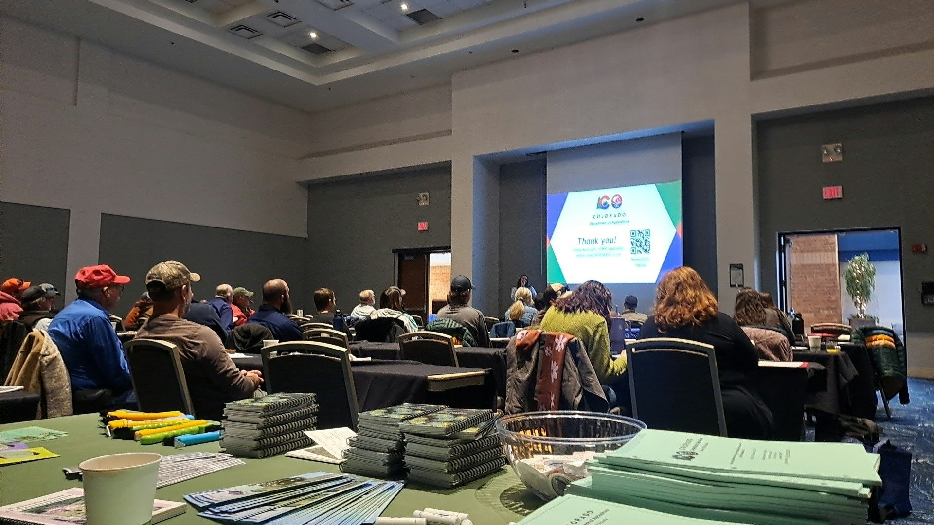 Audience in a conference room with a presentation on a screen; papers and highlighters on a table.