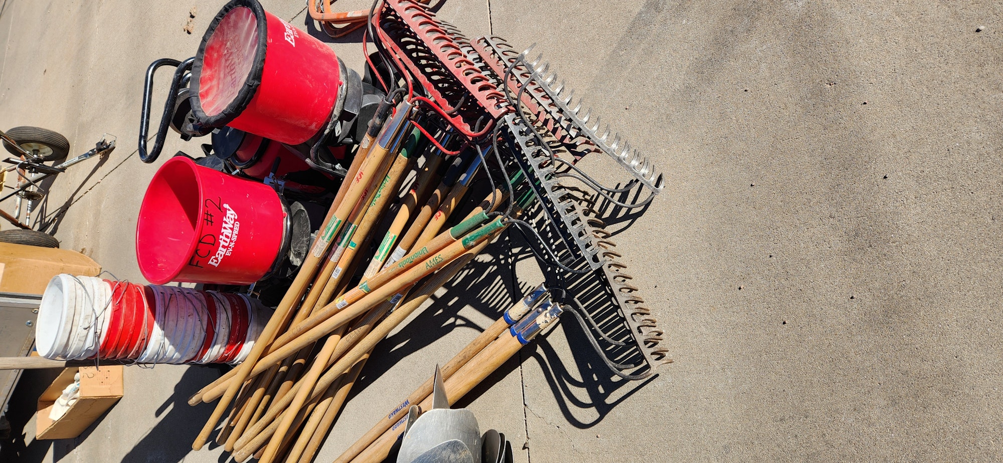 Assorted garden tools and equipment, including rakes, buckets, and a wheelbarrow, laid out on concrete.