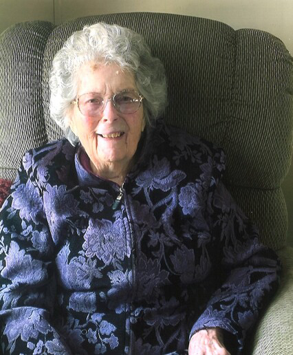 An elderly person with glasses, smiling, sitting in a chair, wearing a floral patterned jacket.