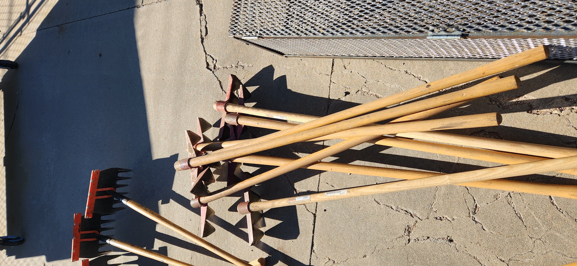 Garden tools with wooden handles and metal parts, including rakes and hoes, laid out on a concrete surface.