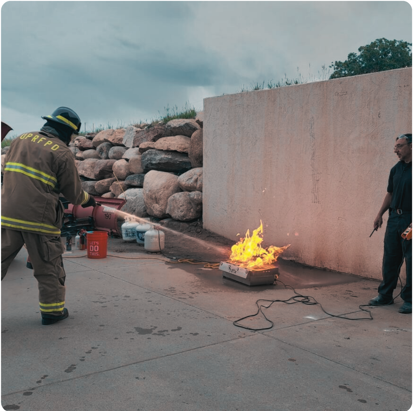 Firefighter using extinguisher on fire during training, observer watches.