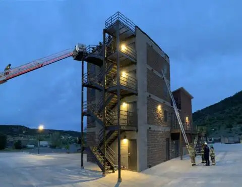 Firefighters training at a multi-story fire drill tower at dusk with a fire truck ladder extended towards the building.