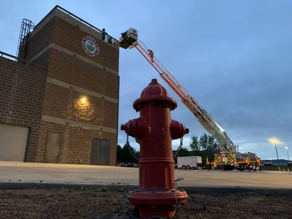 Fire hydrant in the foreground, fire truck with extended ladder, and building in the background at dusk.
