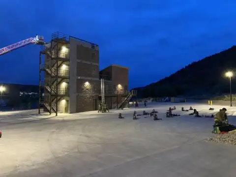 A training facility with a multi-story structure, crane, and people on a concrete pad, likely for firefighting or rescue exercises.