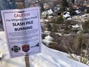 Sign stating "Caution: Slash Pile Burning," snowy landscape and houses in the background.