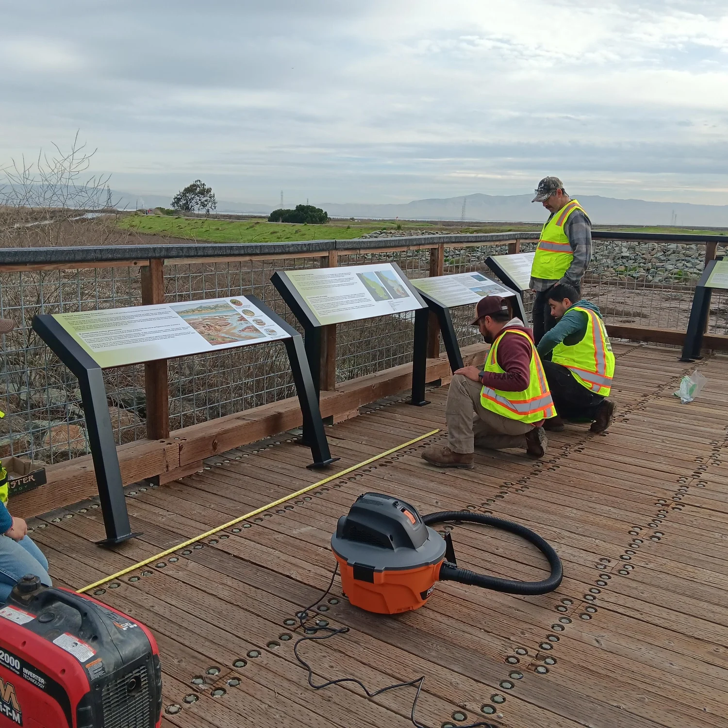 Workers in high-visibility vests are installing informational signs on a wooden pathway near a natural landscape.