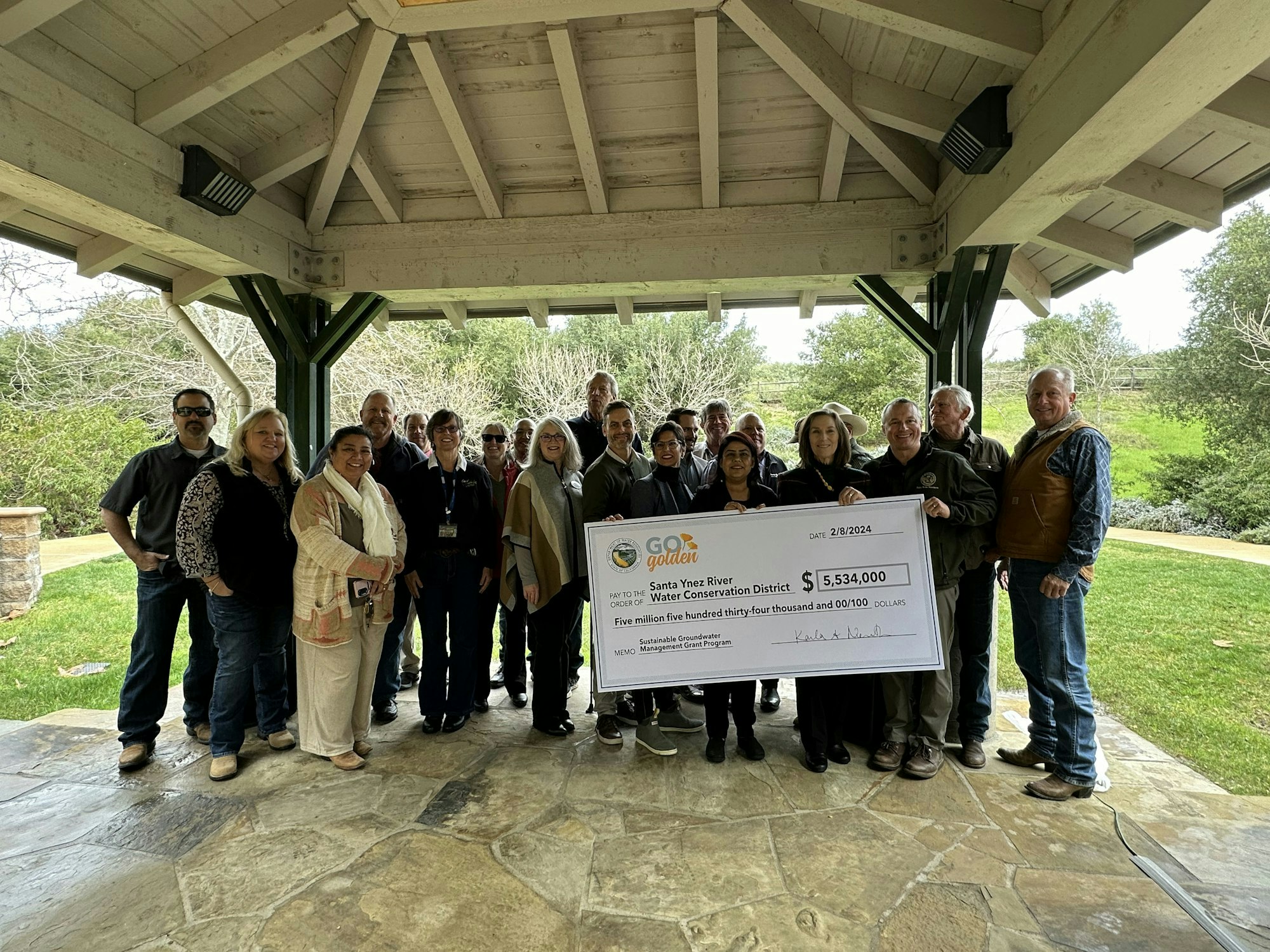 Group of people smiling and holding a large ceremonial check in an outdoor pavilion.