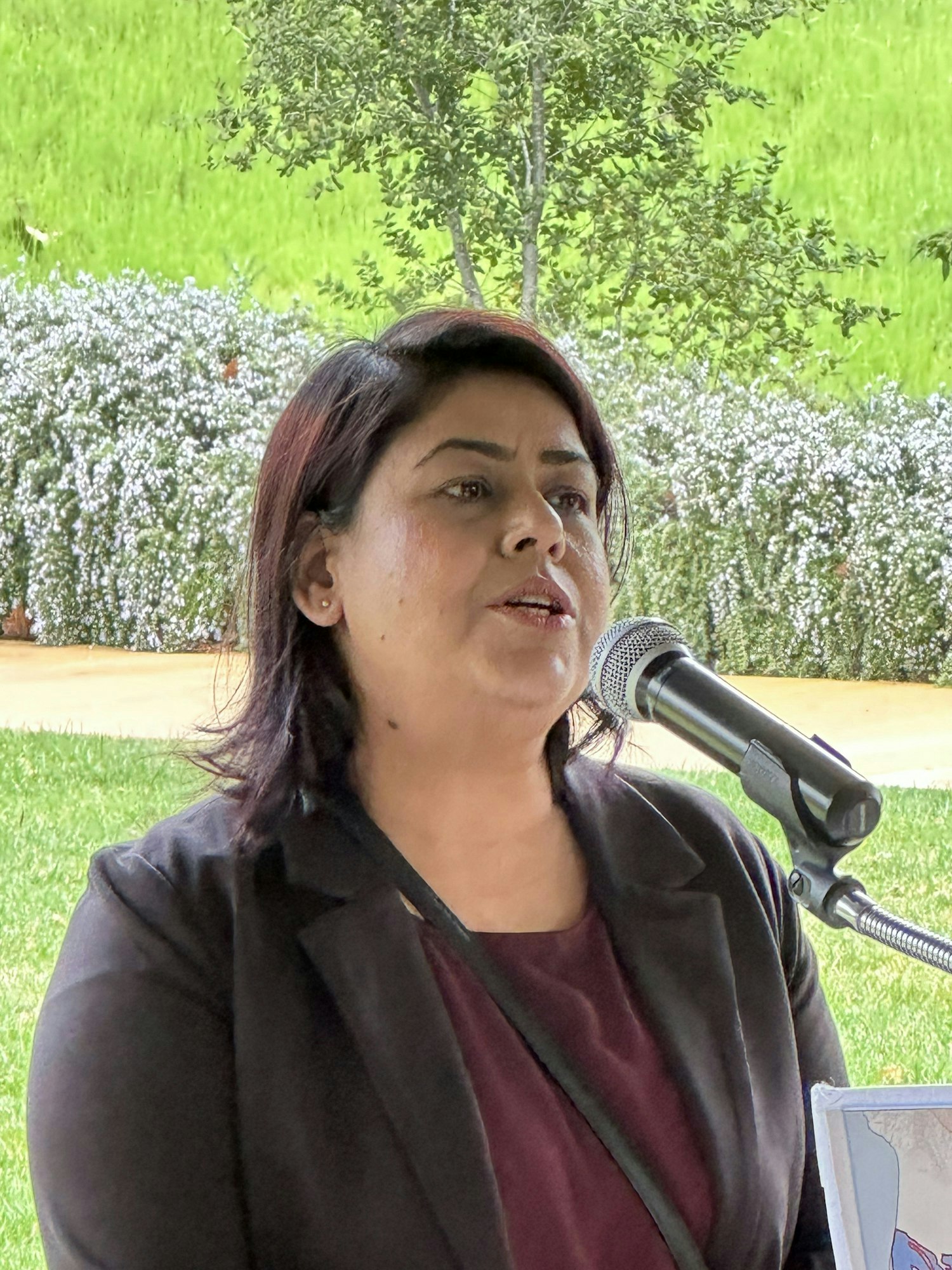 A woman speaking into a microphone, possibly at an outdoor event, with greenery in the background.