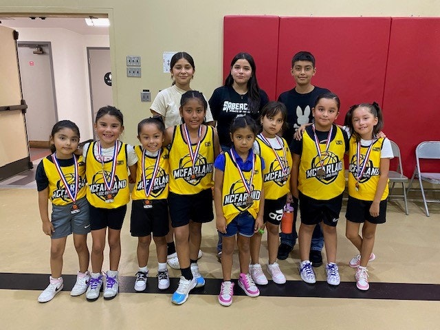 A group of children with medals and two coaches, possibly a youth sports team, standing indoors.