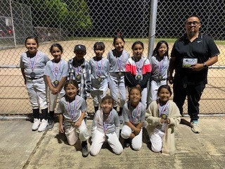 A group of kids in sports uniforms and medals posing with an adult in front of a fence and baseball field at night.