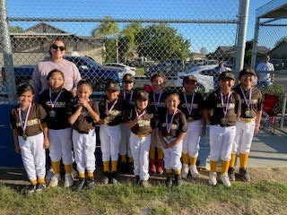 A group of kids in baseball uniforms with medals, posing with an adult in front of a fence.