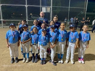 A youth baseball team poses with medals and a coach at a field, wearing blue uniforms and smiling.
