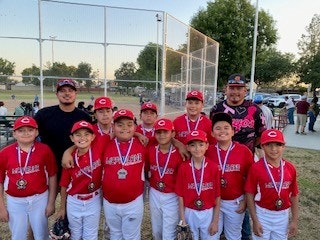 A youth baseball team in red uniforms poses with medals, standing in front of coaches near a baseball field.