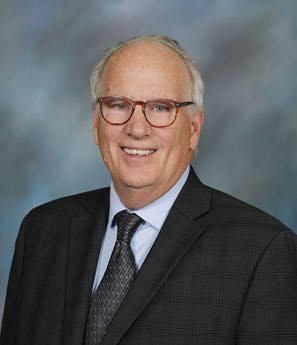 A man in a suit, wearing glasses, smiling in front of a cloudy background.