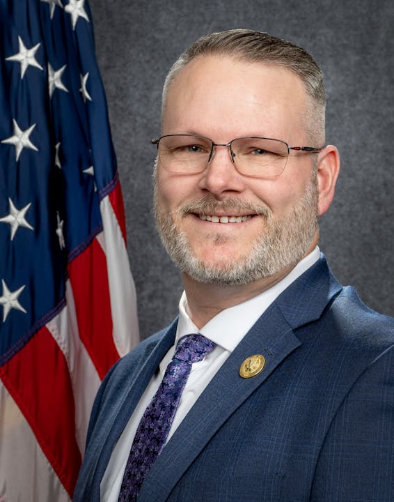 A person in a suit with glasses stands beside an American flag.