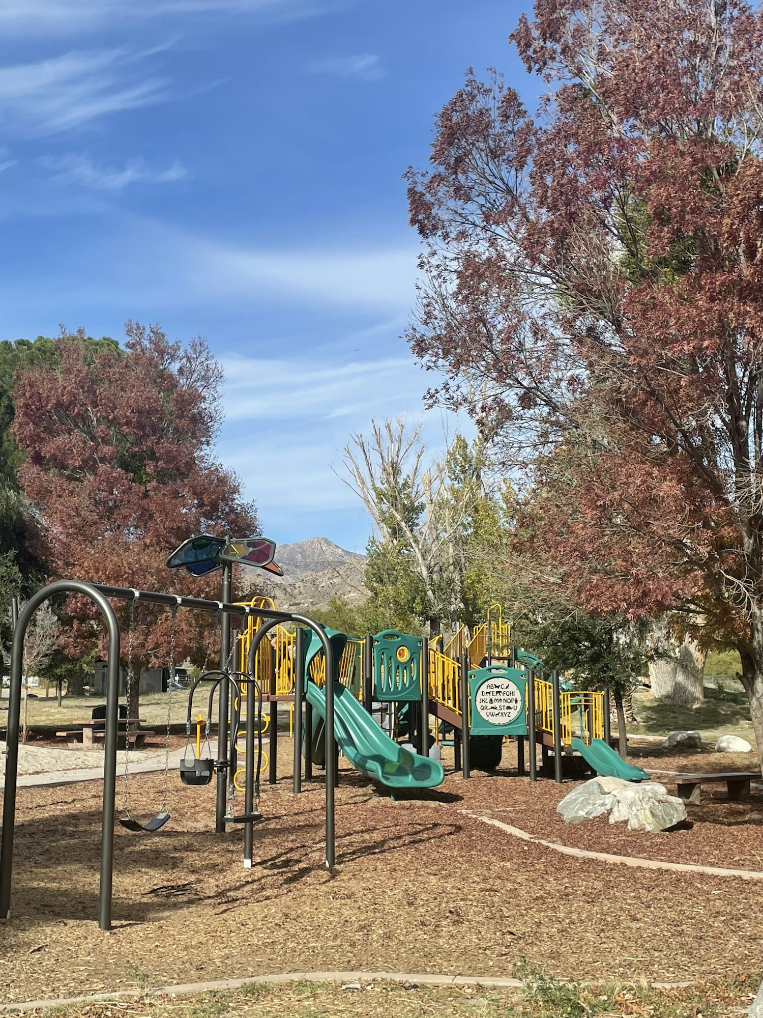 A playground with colorful equipment, trees with autumn leaves, and a blue sky with wispy clouds. (Image is rotated 90 degrees)