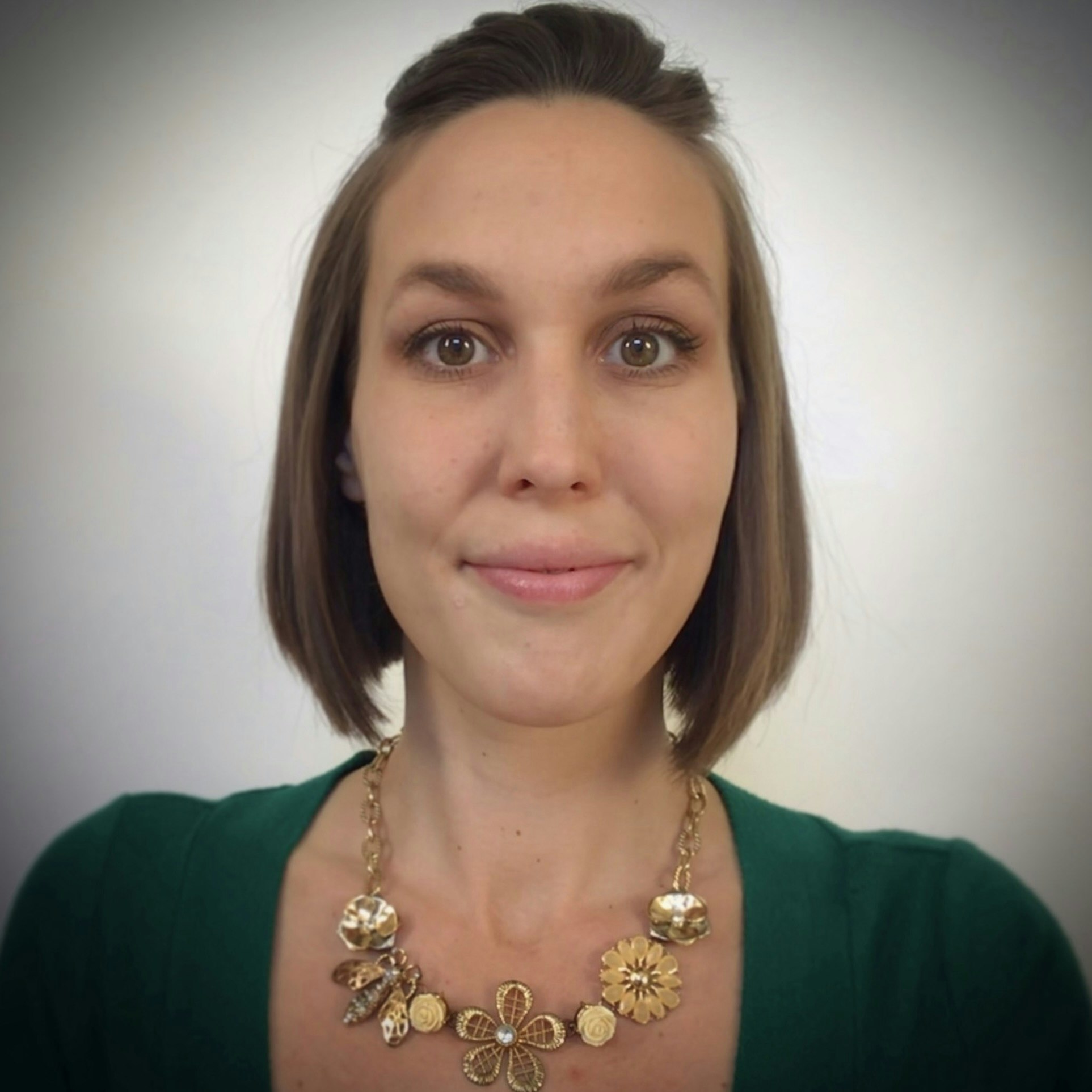 A woman with shoulder-length hair, smiling, wearing a green top and a floral necklace.