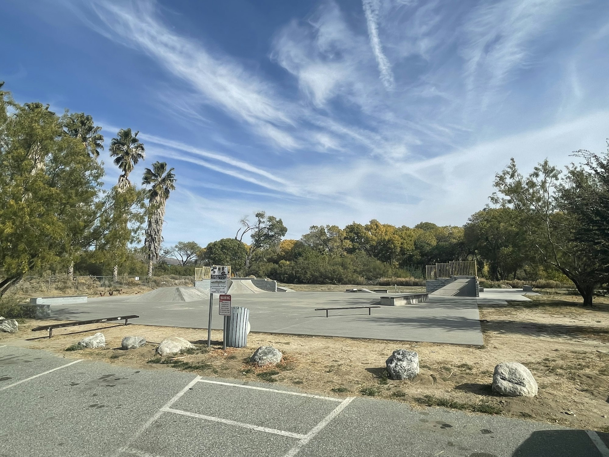 Outdoor skatepark with ramps, blue sky with wispy clouds, palm trees, and greenery around, trash can, bench, and parking space visible.