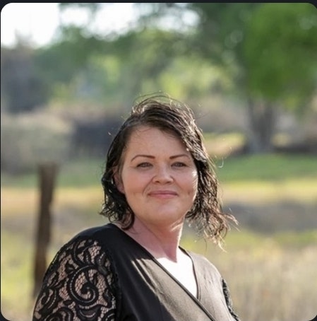 A person with short dark hair standing outdoors, wearing a black lace-sleeved top, with a blurred background of trees and grass.