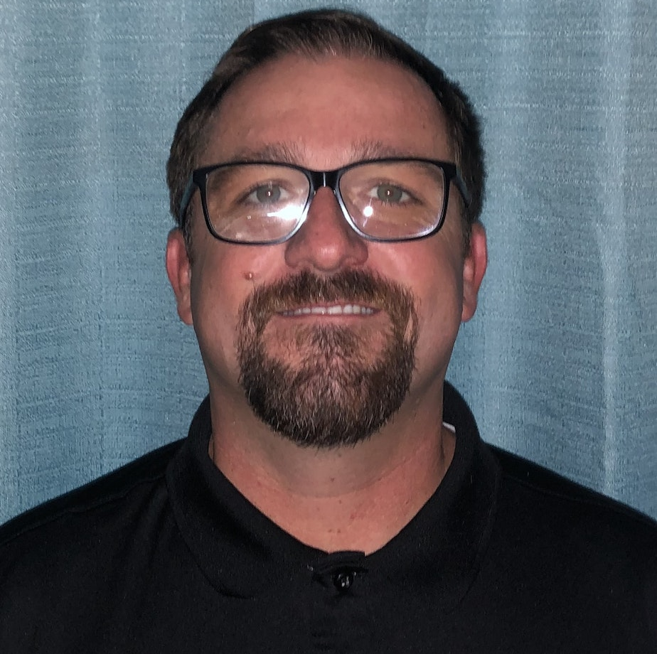 Man with glasses, beard, and black shirt against a blue curtain background.