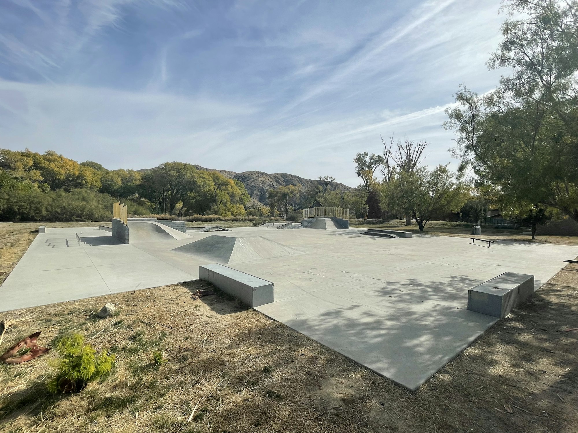 Outdoor skatepark with ramps and railings, surrounded by trees and hills under a clear sky.