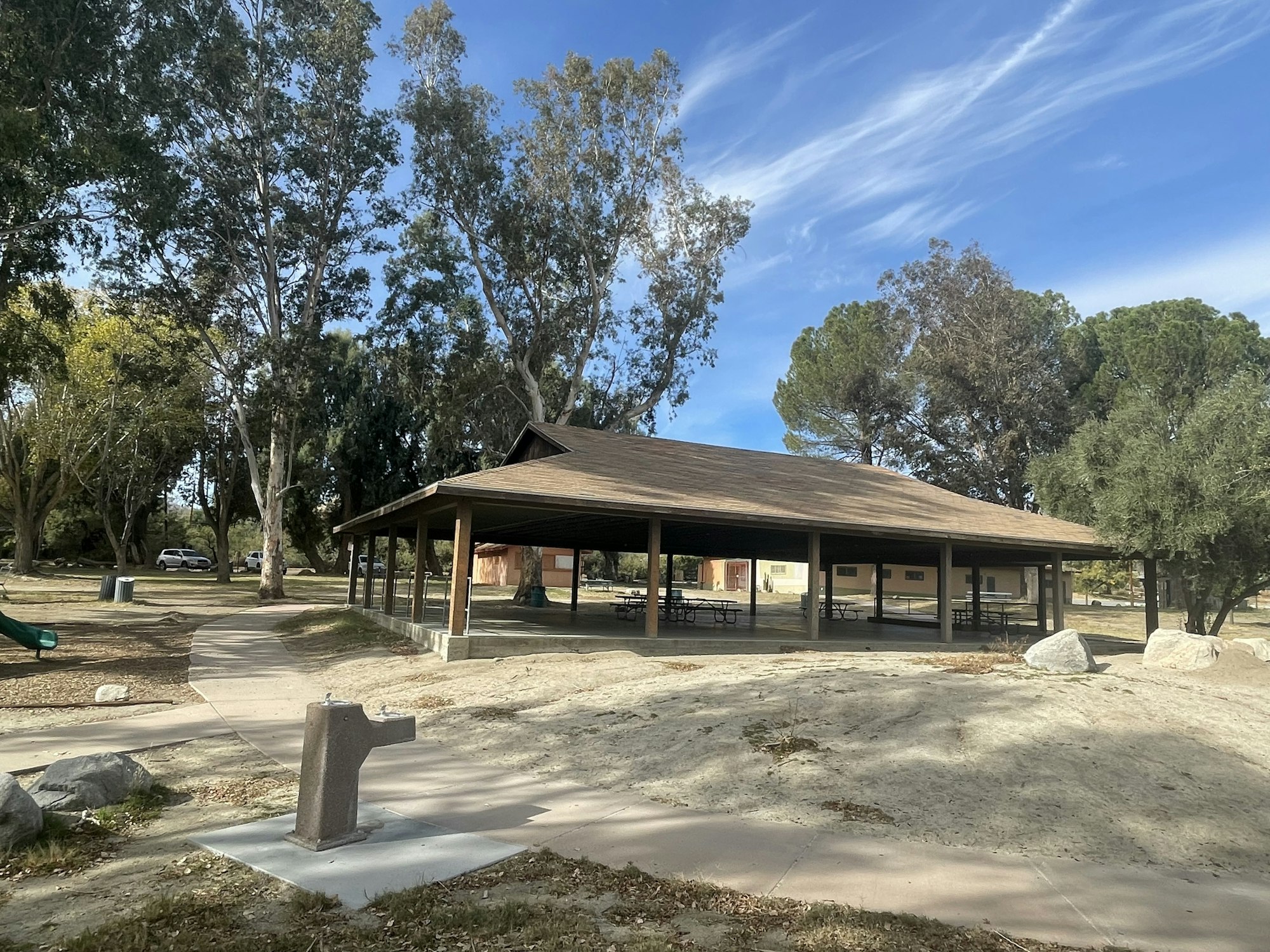 A sunny park scene with a picnic pavilion, tables, trees, a drinking fountain, and a clear blue sky.