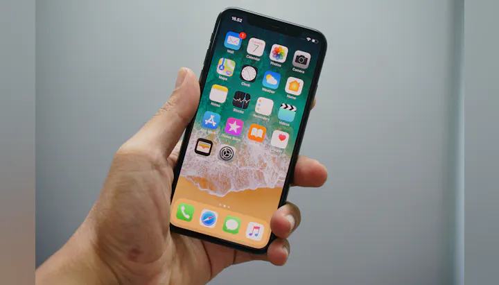 A hand holding a smartphone displaying various app icons on its home screen against a light background.