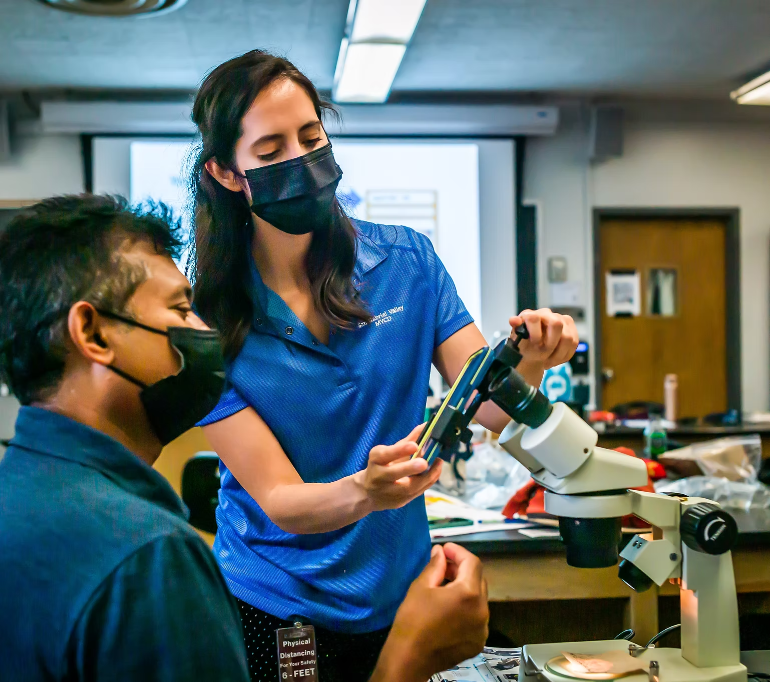 A woman demonstrates a microscope to a man in a classroom setting, both wearing face masks. Educational atmosphere.