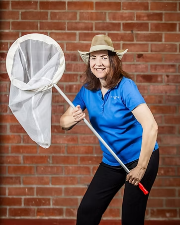 A smiling woman in a blue shirt and hat poses with a butterfly net against a brick wall.