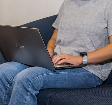 A person sitting on a couch, using a laptop, wearing a gray t-shirt and jeans, and a watch on their wrist.
