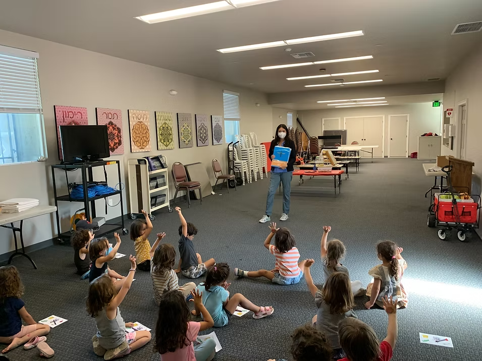 A teacher in a mask addresses a group of children sitting on the floor, raising their hands in a classroom setting.