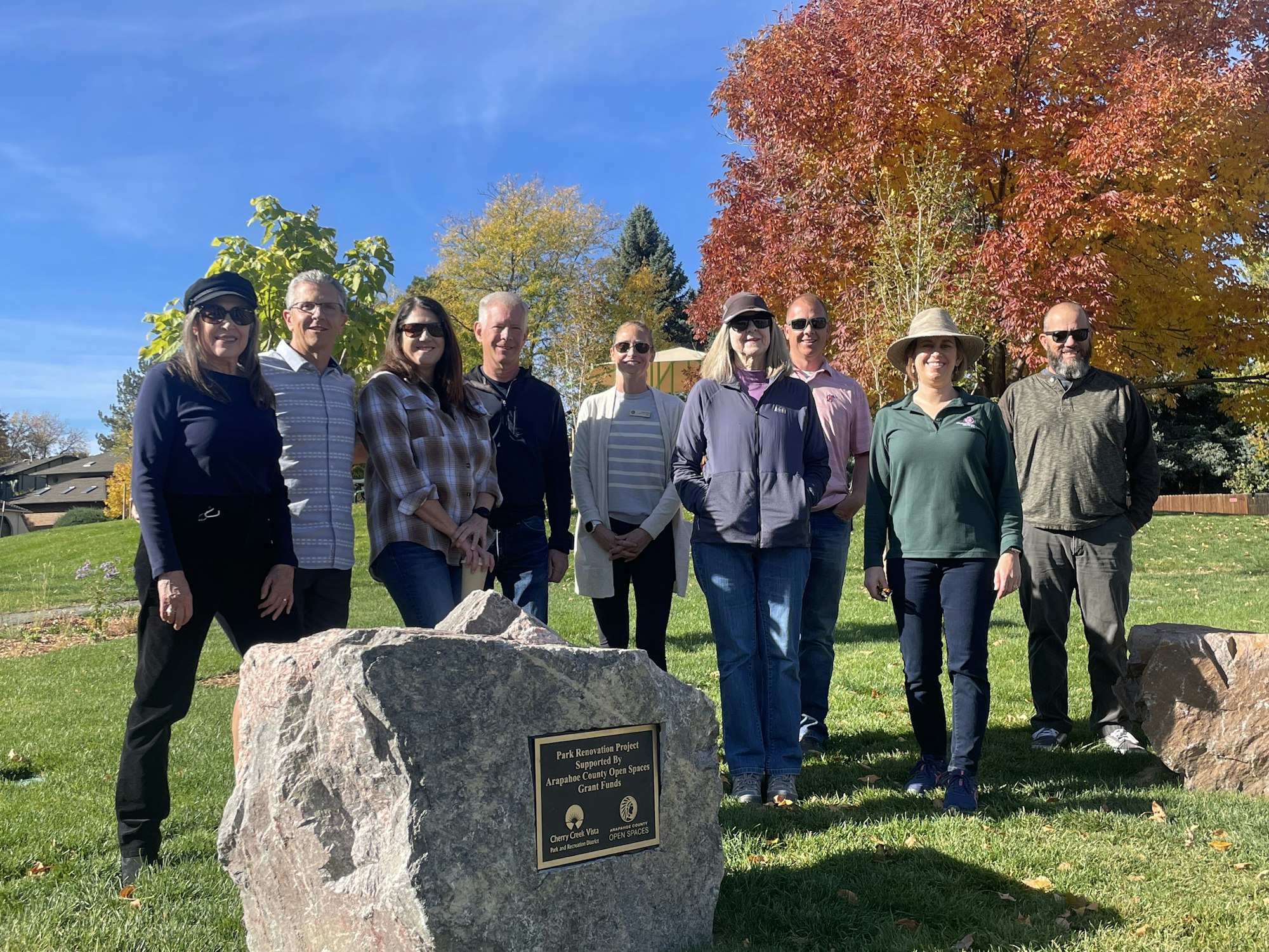A group of people posing behind a commemorative plaque in a park with autumn trees.