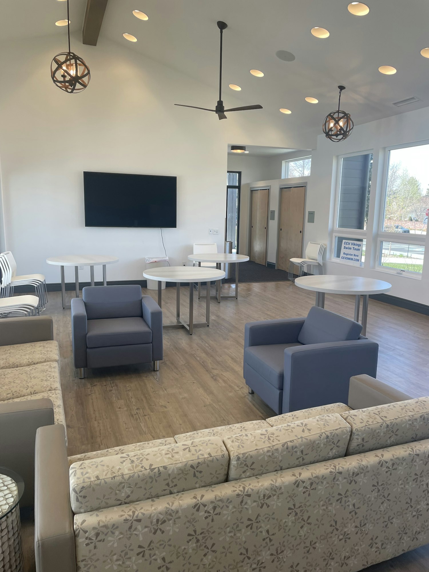 A modern lounge with sofas, armchairs, tables, a TV, ceiling lights, and fans.