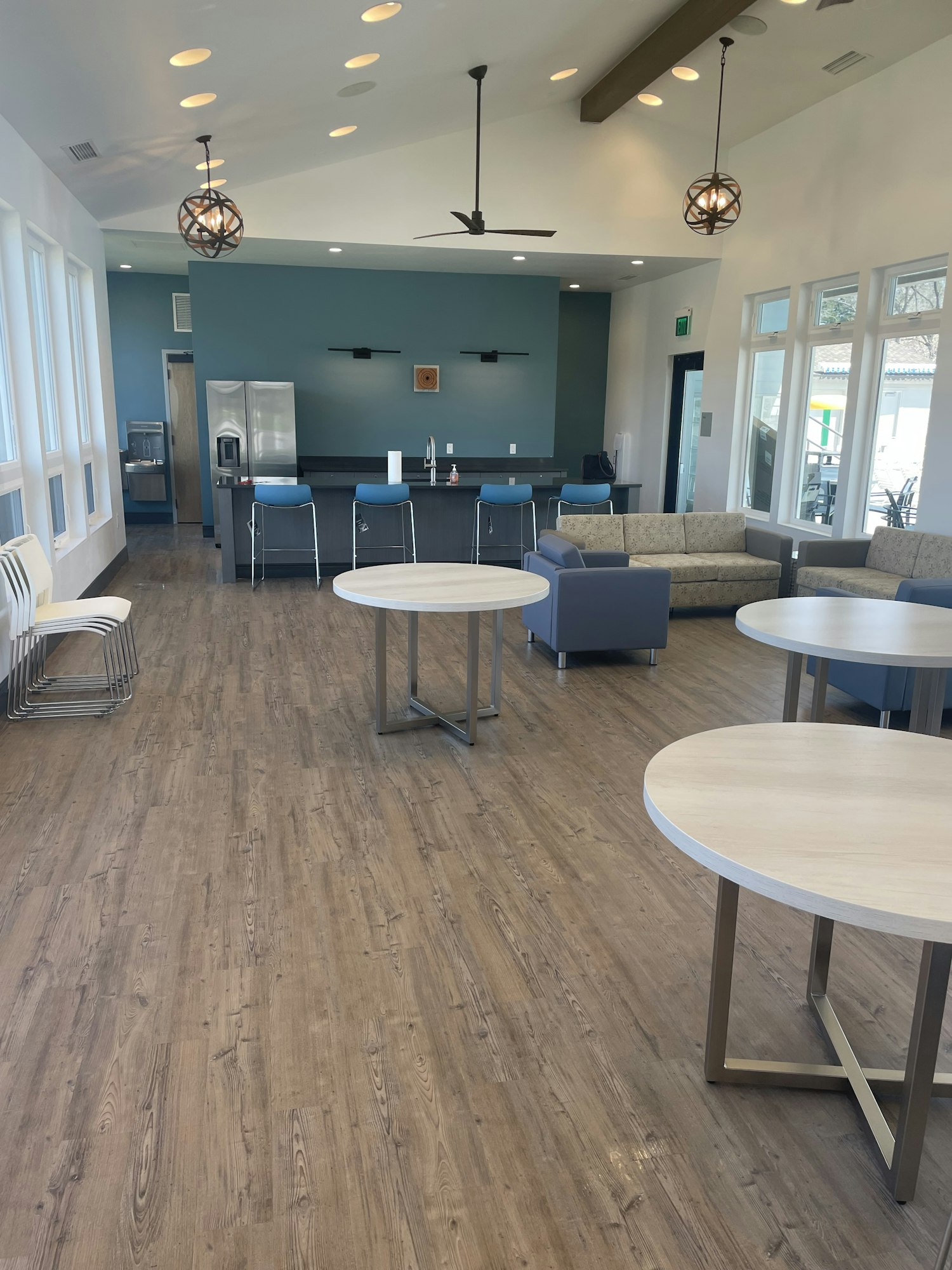 Modern room with wooden floor, round tables, blue chairs, fridge, and windows. Ceiling fans and light fixtures, lounge in the background.