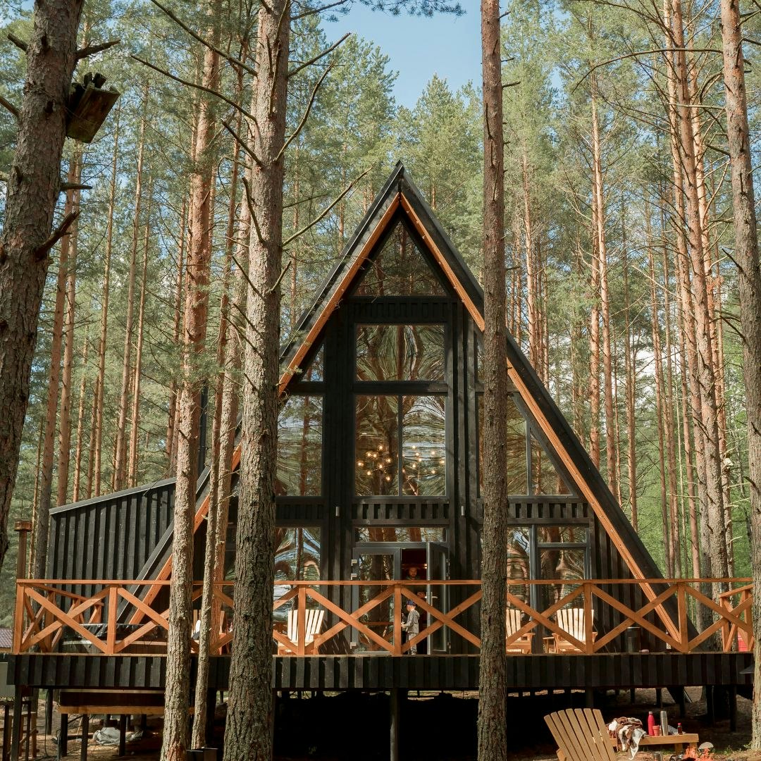 A-frame cabin in a forest, surrounded by tall trees, with a wooden deck and seating area.