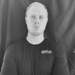 A black and white portrait of a person wearing a dark shirt, standing against a dark background.