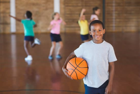 A smiling child holds a basketball in the foreground, with other children playing in the background.