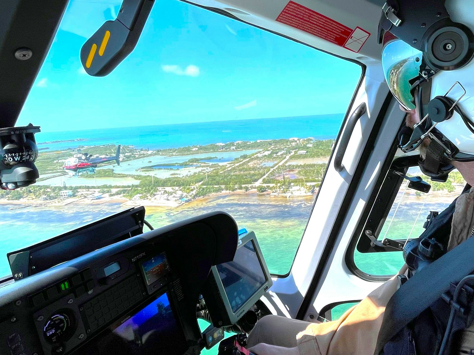 Cockpit view from a helicopter flying over coastal landscape with clear skies and another helicopter visible outside.