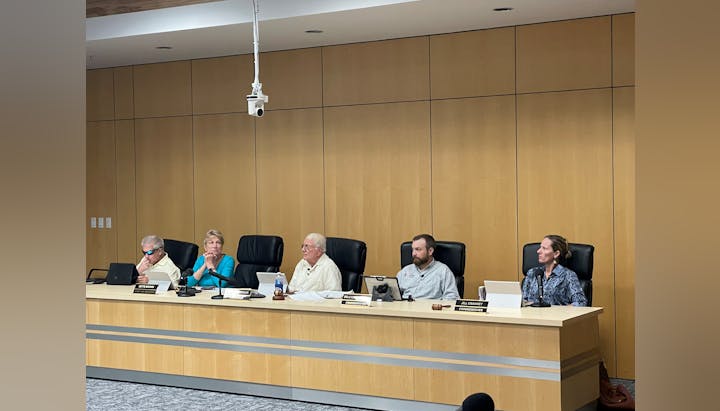 A panel of five people sitting behind a long desk in a conference room with microphones and nameplates.