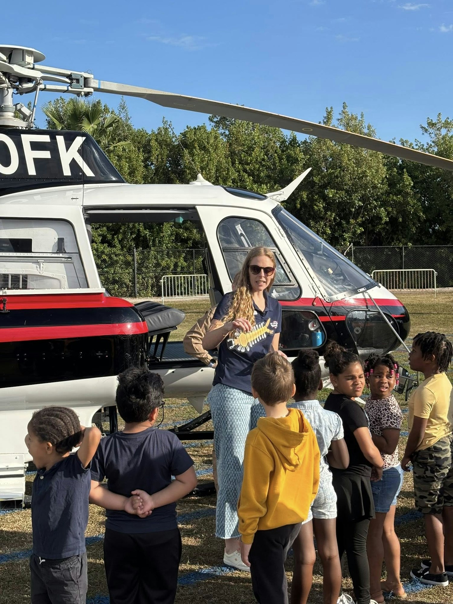 A group of children is gathered around a helicopter, listening to a woman in sunglasses explaining something enthusiastically.