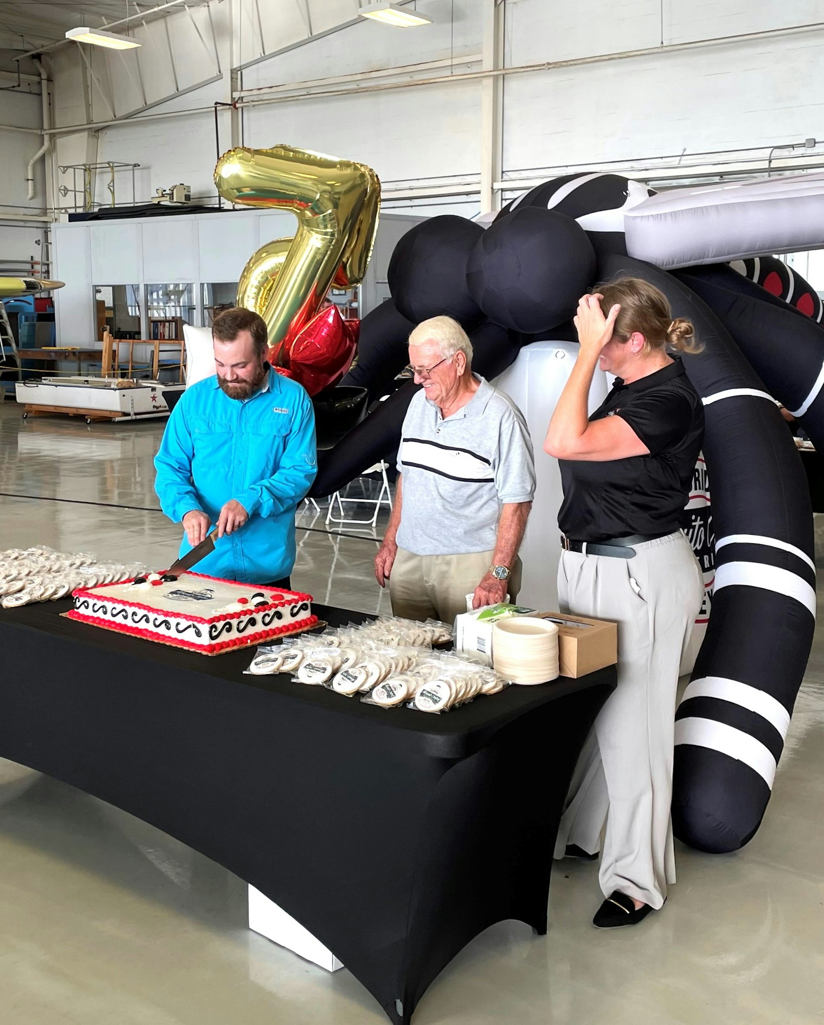 Three people celebrate with a cake, number balloons, and inflatable in a hangar.