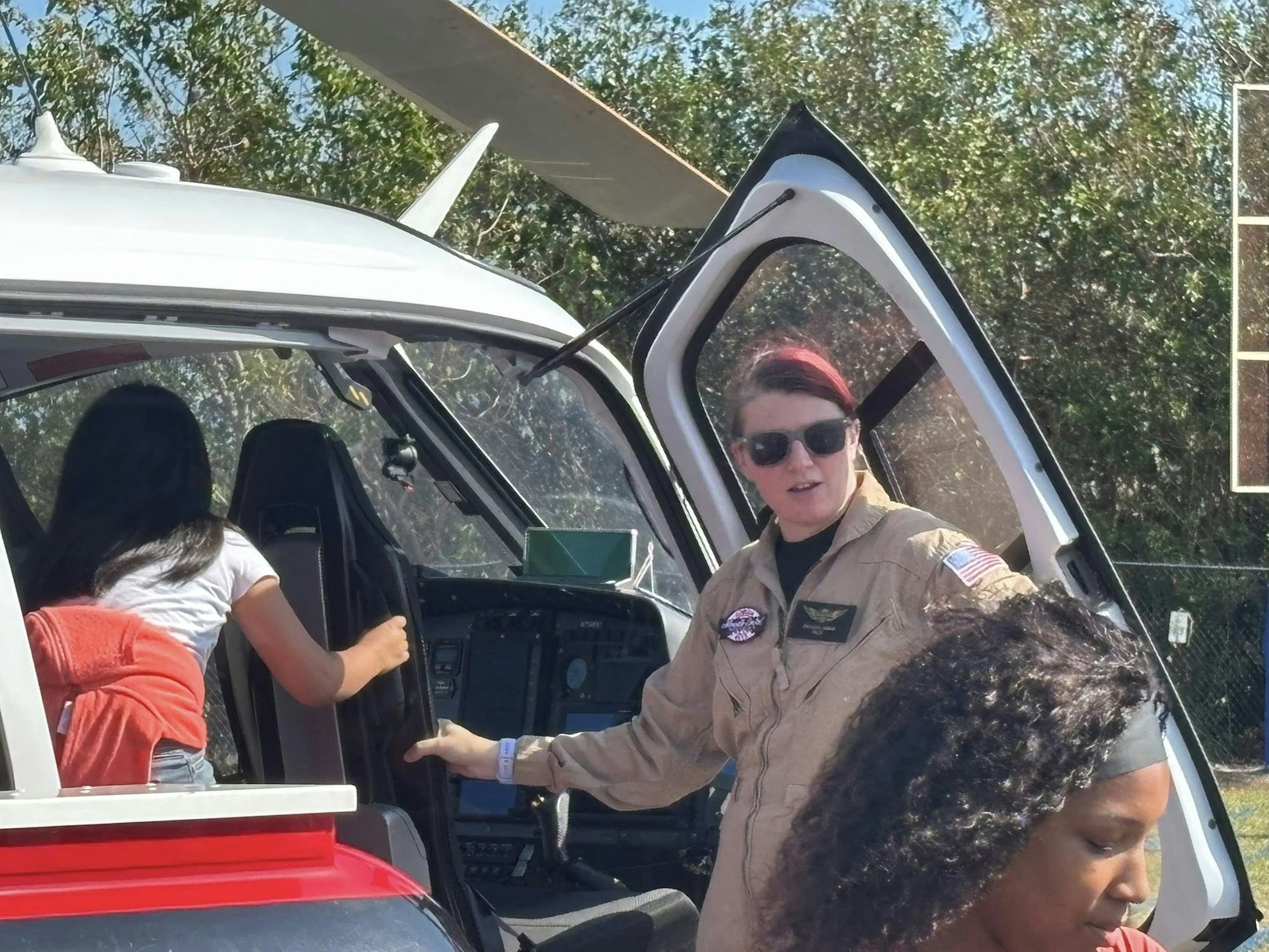 A helicopter scene with a pilot in sunglasses and a female passenger entering the cockpit, with another person nearby.