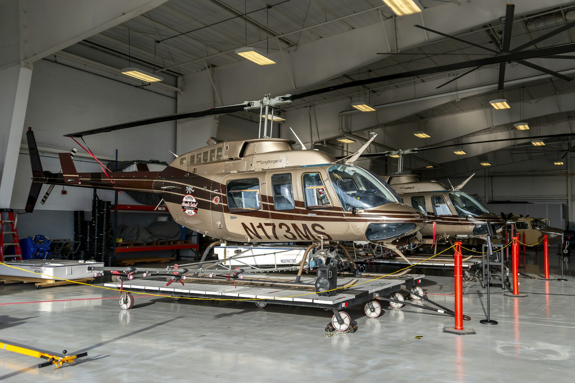 A helicopter inside a hangar, surrounded by maintenance equipment and another helicopter in the background.