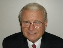 A man with gray hair and glasses, wearing a suit, appears to be posing for a photo against a plain background.