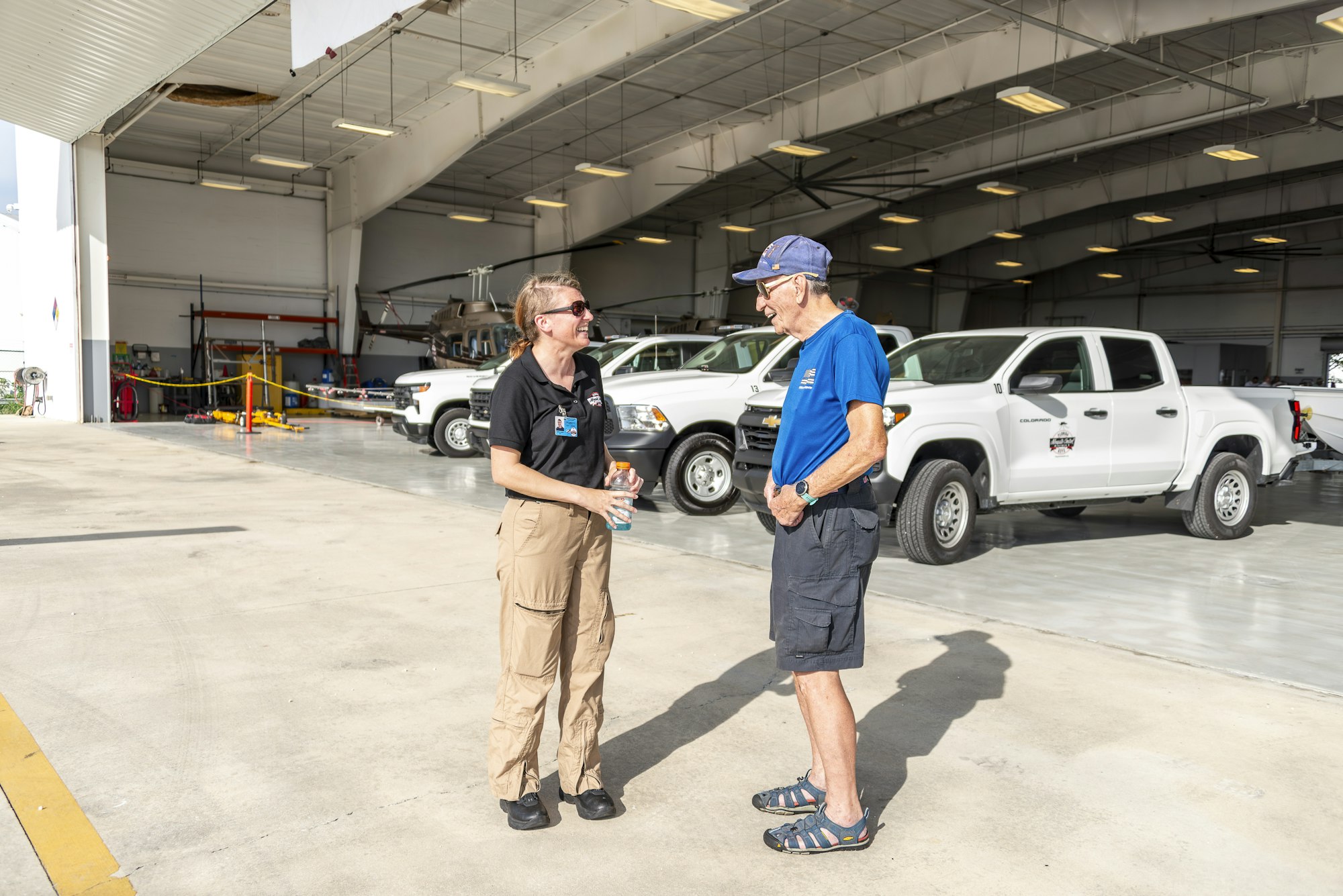 Two people talking in an aircraft hangar with parked white trucks and a helicopter in the background.