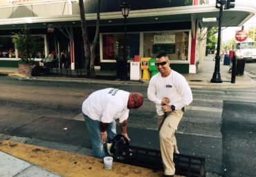 Two men working on a bench on a city sidewalk. One is standing and observing while the other is crouching and drilling.