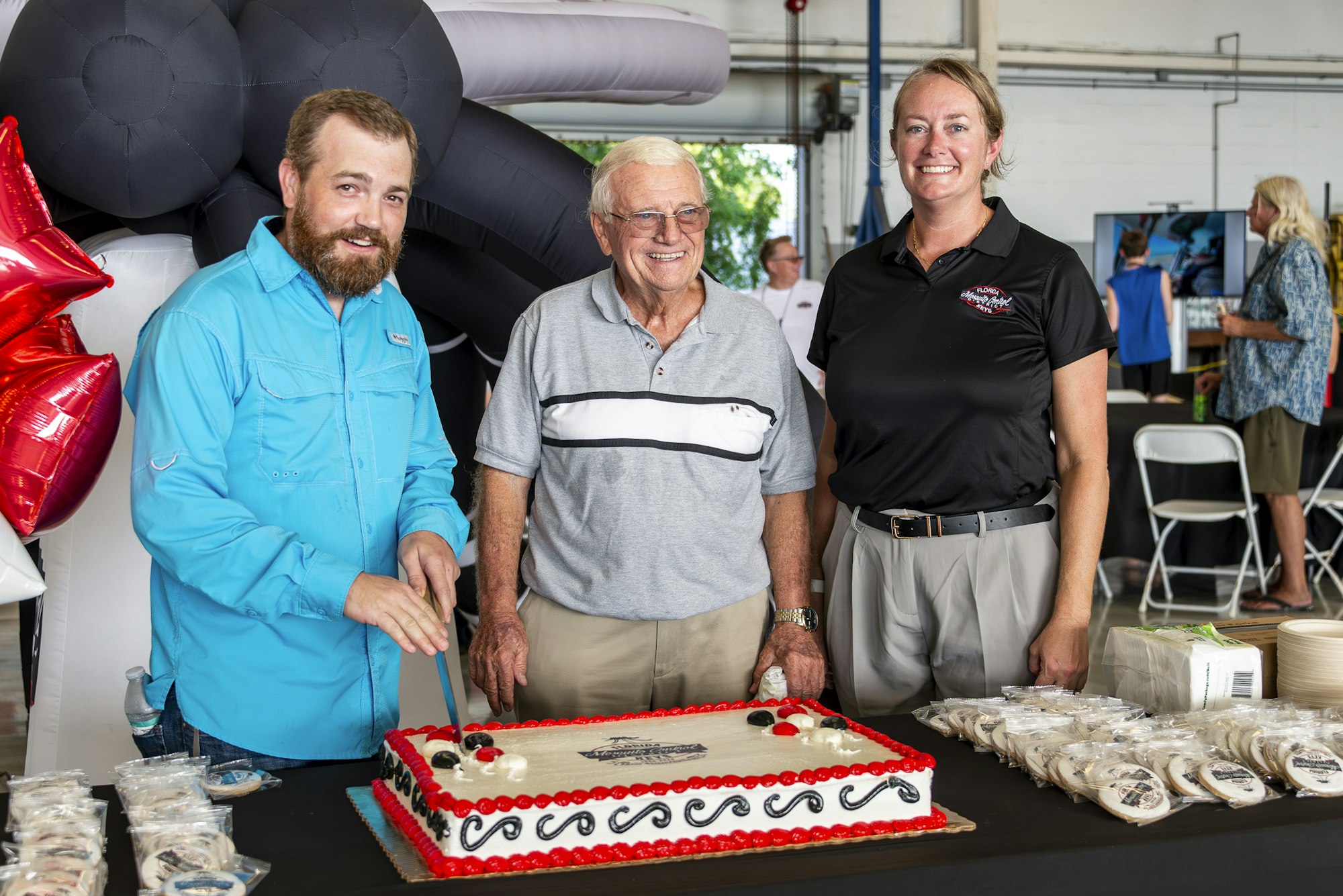 Three people smiling in front of a decorated cake; one is about to slice it. There are balloons in the background.