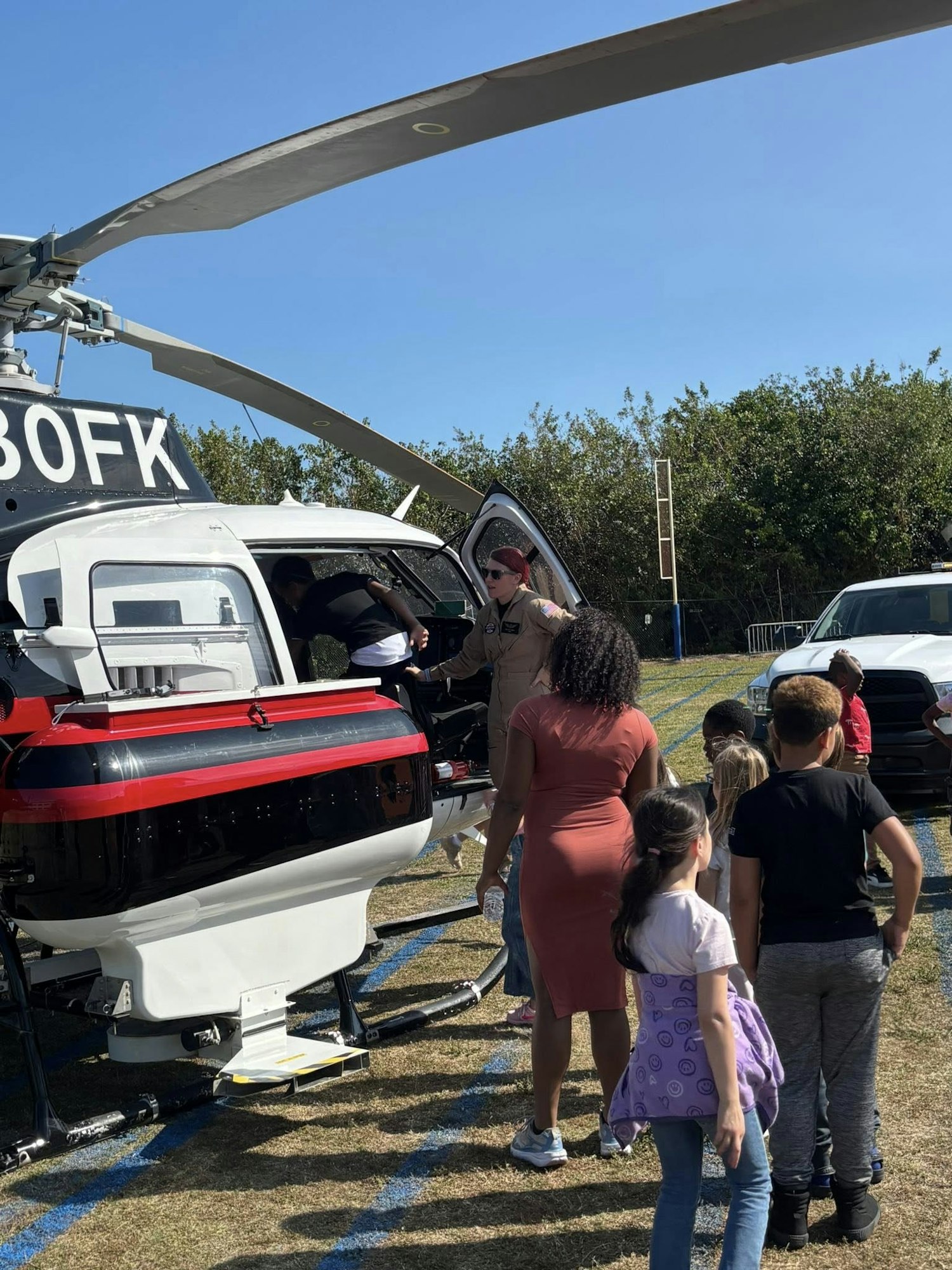 A group of children gathers around a helicopter, with an adult assisting them as they explore the aircraft on a sunny day.