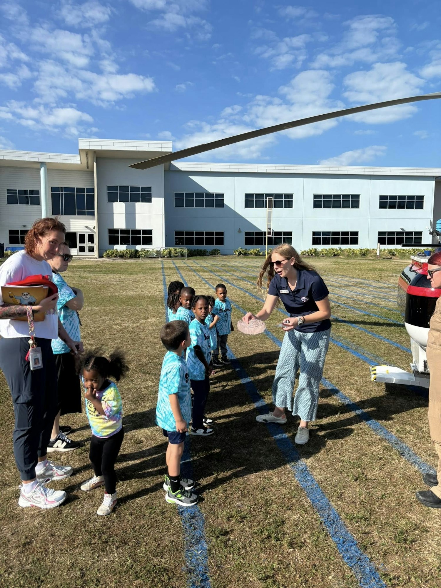 Children in colorful shirts line up outside a building, engaging with an adult near a helicopter on a sunny day.